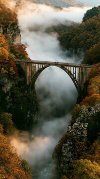 a bridge spanning a gorge, bathed in the warm hues of autumn. The bridge, with its arches and pillars, is the focal point, surrounded by a dense forest of trees displaying a mix of vibrant red.