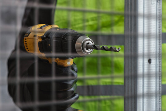 Worker holding yellow cordless electric drill driving screw into metal stud frame behind wire mesh fence. Drywall installation, interior renovation, construction worker and power tool concept.