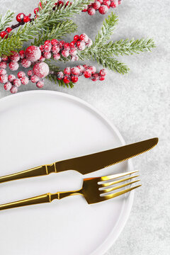 Festive holiday table setting with golden cutlery arranged on white plate with showy Christmas tree branch with red holly berry on light gray textured background. Knife and fork close up, macro