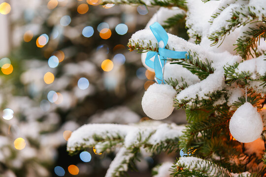 Snow covered Christmas tree branches decorated with white ornaments and a blue ribbon on glowing fairy lights colorful bokeh background. Cozy winter holiday atmosphere.