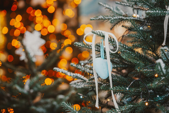 Snow covered Christmas tree branches decorated with heart shape ornament and a blue ribbon on glowing fairy lights colorful bokeh background. Cozy winter holiday atmosphere.