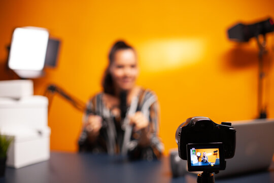 Camera and flip-out screen are in focus while a blurred woman gestures during a studio recording, capturing behind-the-scenes vlog and digital content creation.