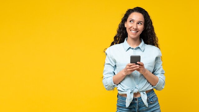 Smiling Young Woman With Dark Hair And Light Skin Holding Smartphone While Looking Upjoyfully At Studio Background. She Engaged In Communication, Expressing Positivity And Connection.