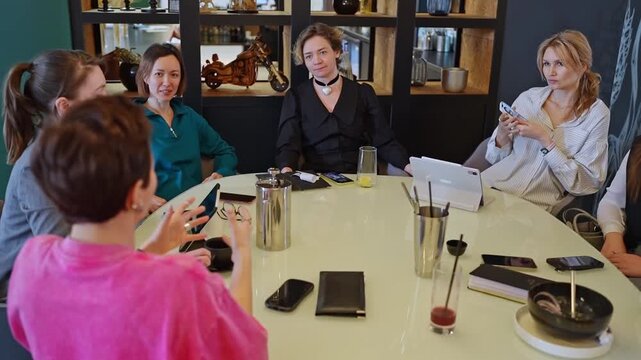 Group of female colleagues enjoying lunch and conversation around round table in modern restaurant, warm lighting, glass partition with frosted pattern, casual professional dining atmosphere