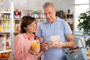 Fototapeta premium Elderly couple examines packs of cereal in store, chooses and buys instant porridge. Buyer is thinking about choosing variety of flakes