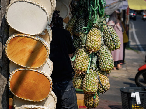 Fresh pineapples and oval wooden trays tied in green raffia string hanging at a traditional street fruit stall.