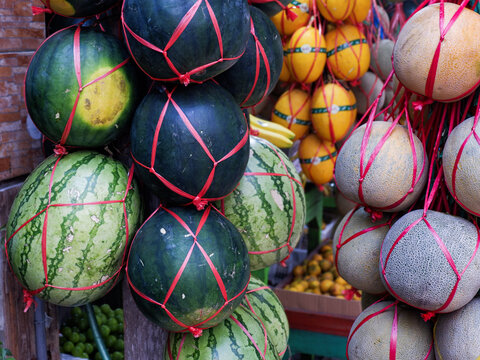 Fresh watermelons and cantaloupes tied in red plastic raffia string mesh hanging at a traditional fruit market stall.