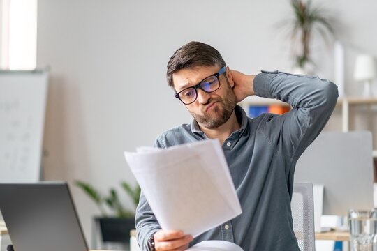 Worried Caucasian Man In His 30s Sitting At Desk In Modern Office, Reading Important Documents With Concerned Expression. Feeling Overwhelmed While Working On Business Task At Workplace.