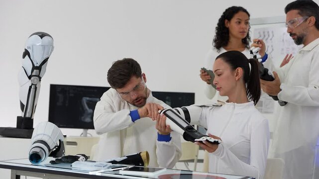 Wide view of a bionics workbench with robot leg models as an engineer secures a prosthetic forearm and a woman holds the black hand while colleagues watch from behind