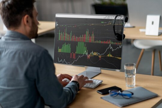 Focused Man Analyzing Stock Market Data On Computer Screen While Working In Bright Modern Office Environment. Demonstrating Professional Engagement With Financial Information, Business Concept.