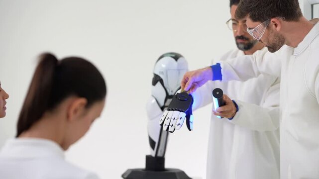 Close-up of an engineer pointing with a pen at a robotic hand mounted on a white robot arm stand and holding a blue UV torch while a woman with a ponytail watches