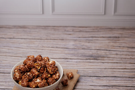 Caramel-coated popcorn in a white bowl sits on burlap. Wooden table and panelled wall frame the scene. Warm tones evoke cozy, comforting nostalgia. Ideal for food blogs, recipe ads
