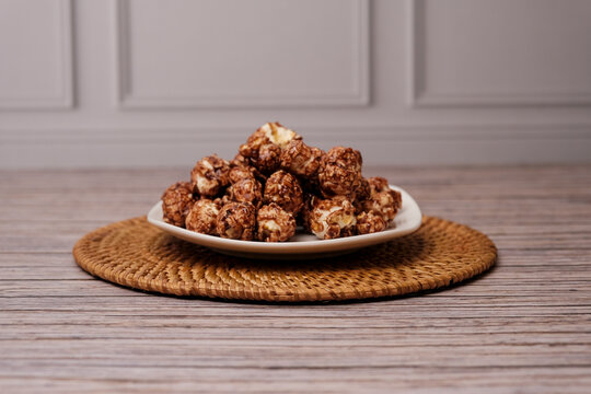 Chocolate popcorn piled high on a white plate. Plate rests on a woven, round mat. Wooden surface beneath adds rustic charm. Sweet, crunchy snack looks deliciously inviting