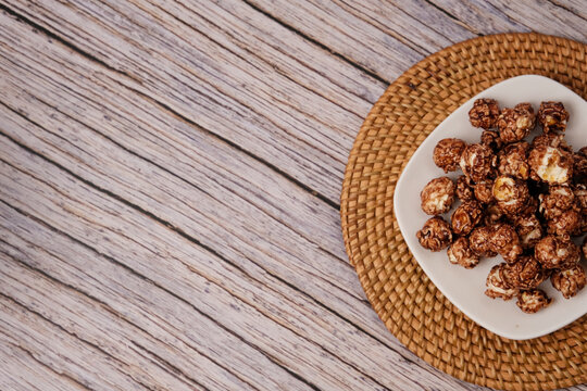 Chocolate popcorn piled high on a white plate. Plate rests on a woven, round mat. Wooden surface beneath adds rustic charm. Sweet, crunchy snack looks deliciously inviting