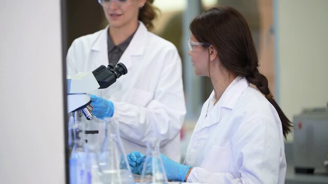 Two female scientists share a microscope, one with a ponytail peering in while a brunette colleague with a braid mixes solutions in flasks, then a third looks on