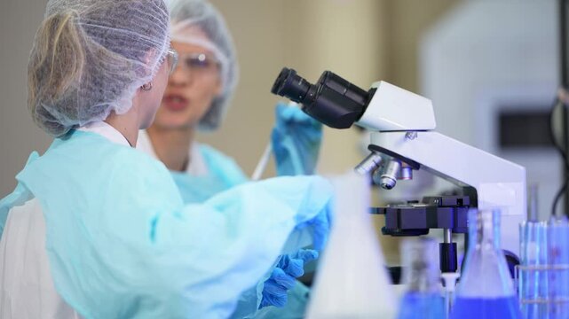 Two women in light blue gowns and hairnets work side by side at a bench, one smiling while another uses a pipette, microscope and flasks of blue liquid nearby