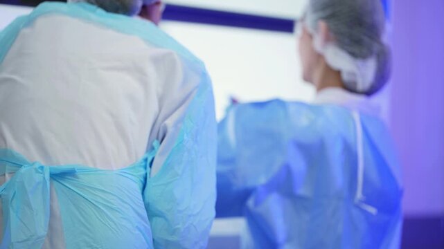 Scientist in a blue gown and hairnet raises a test tube in a glowing laboratory hood, a colleague standing in the blurred foreground, rack focus to the tube