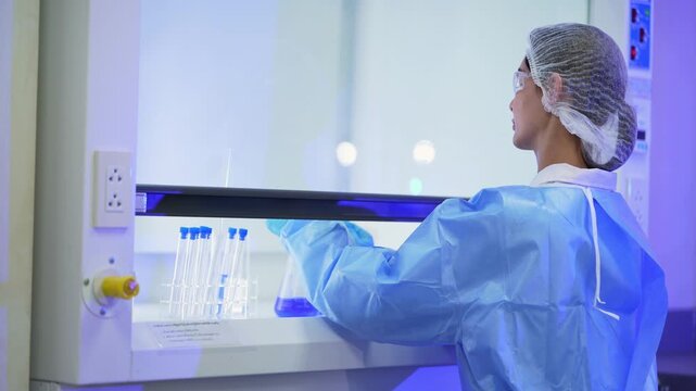 Wider shot of a female scientist in a blue gown and hairnet using a fume hood, raising the sash and handling a blue flask and tubes with electrical sockets beside