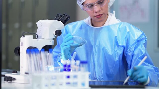 Scientist in a blue gown turns and inspects a petri dish while writing notes with a stylus on a tablet next to a microscope and rows of purple-capped tubes