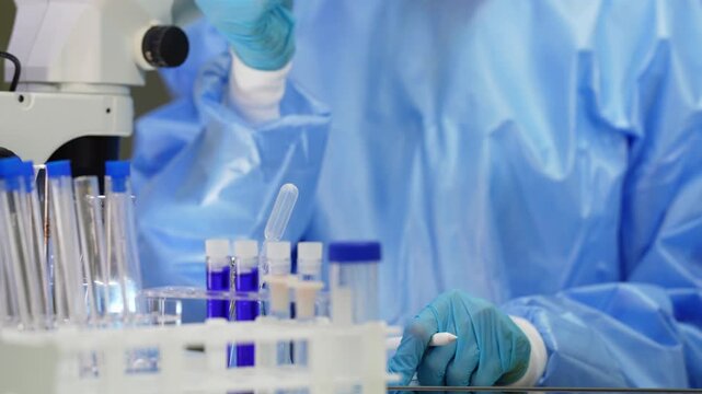 Close-up of blue-gloved hands in a disposable gown lifting a sample tube above a rack of blue-capped test tubes and purple vials, no face in view
