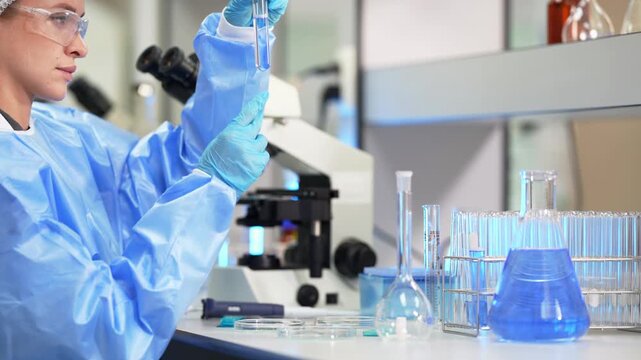Side view of a woman in a blue gown holding a glass pipette upright above a microscope, with a large flask of blue liquid and racks of test tubes on the bench