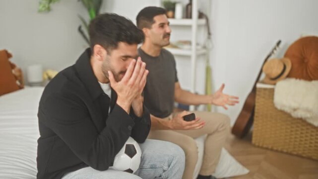 Two men on a bed, one holding a soccer ball and the other holding a tv remote, hands raised and facepalm gestures in a residential building; frustration rivalry friendship.