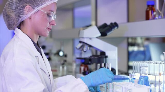 Woman in white lab coat transfers liquid into a test tube with a dropper, then picks up a micropipette to continue sample preparation at the bench