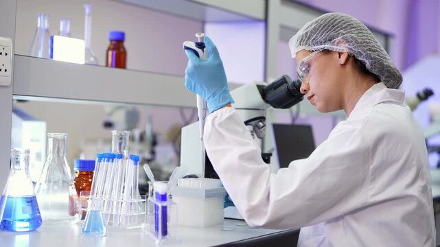 Woman in white lab coat looks into a microscope while holding a pipette in her raised blue glove, with tubes, flasks and reagent bottles filling the bench