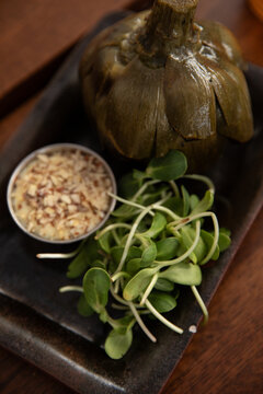 oiled roasted artichoke served with microgreens and dipping sauce with chopped nuts on a restaurant table, selective focus