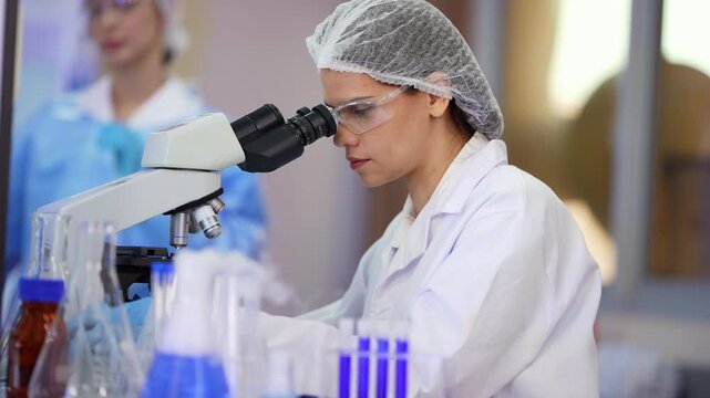 Female lab technician in white coat and hairnet looks through a microscope, then handles a sample in front of blue flasks while a colleague in a blue gown works behind her