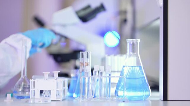 Close-up of a blue-gloved hand holding a pipette above a volumetric flask, with test tubes, a conical flask of light blue liquid and a blurred microscope behind