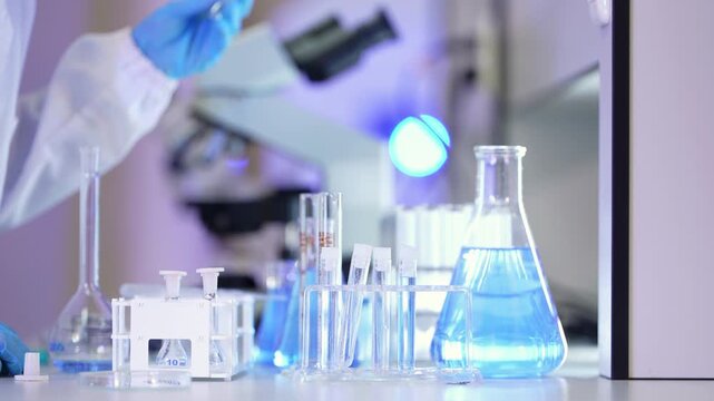 Still close-up of laboratory glassware, a flask of pale blue liquid, test tubes and a volumetric flask in front of a softly blurred microscope and glowing lamp
