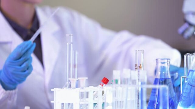Low close-up of gloved hands transferring blue liquid from a pipette into a flask above a rack of test tubes, with a scientist in a lab coat partly out of frame