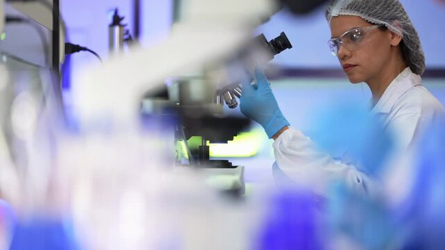 Blurred scientist in a blue protective gown pours a sample into a small flask behind racks of blue test tubes and a glowing microscope, shallow focus