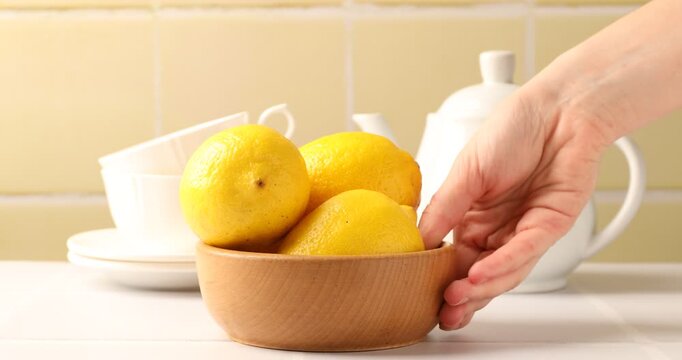 Woman putting bowl with lemons onto white table against yellow tiled wall, closeup