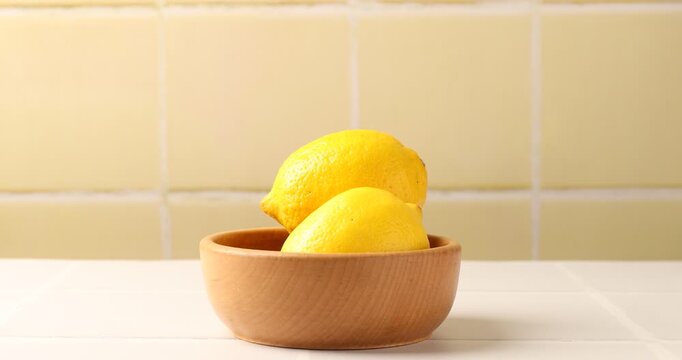 Woman taking lemon from wooden bowl at table against yellow tiled wall, closeup