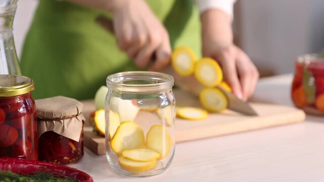 Making pickles. Woman cutting zucchini and putting it into glass jar at white wooden table indoors, closeup