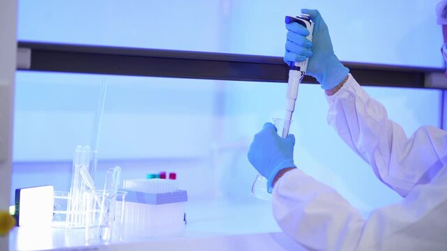 Close crop of a gloved hand holding a pipette high and lowering it toward a flask in a bright blue-lit lab, the scientist's face partly out of frame