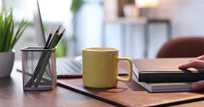 Woman putting yellow mug of coffee and pen onto wooden table indoors, closeup
