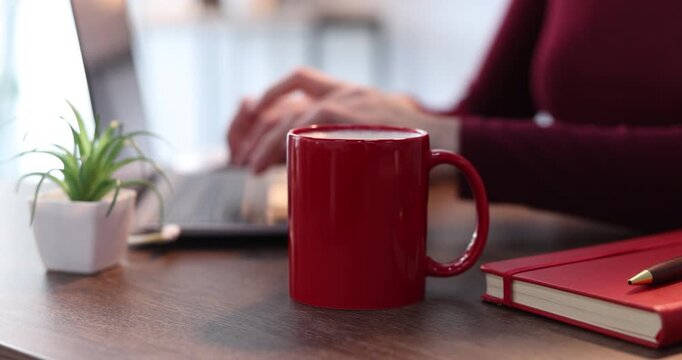 Woman working with laptop and taking mug of coffee at wooden table indoors, closeup