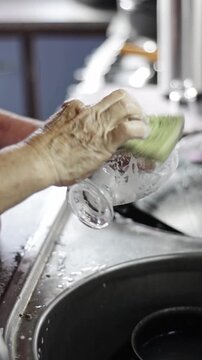 Female hands scrubbing a glass with a sponge in a kitchen sink, close-up of senior woman washing dishes, handheld camera