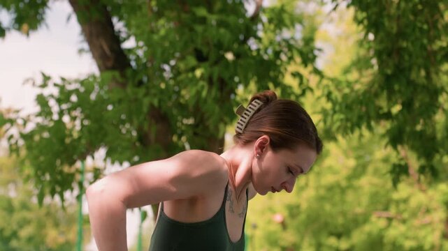 Close up of woman performing outdoor exercise on parallel bars in daylight with green foliage background. Woman is wearing athletic clothing and appears focused during physical activity