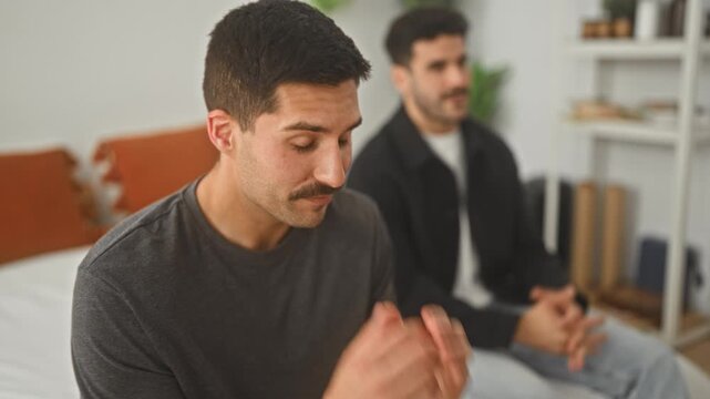 Man holding his face while seated on a bed in a residential building, hands pressing cheeks and temples with a friend nearby; anxiety support compassion.