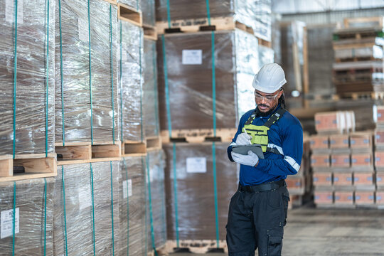 Black worker with a barcode scanner records wrapped goods in navy reflective workwear inside a large timber as part of a smart factory research project on AI, robotics and timber logistics