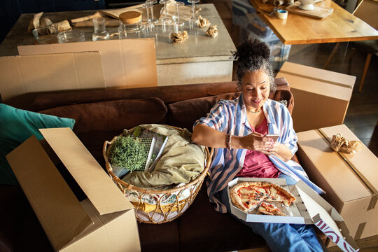 Woman eating pizza and texting while unpacking boxes