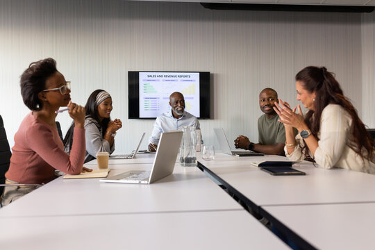 Diverse coworkers discussing sales and revenue reports on wall screen at meeting table with laptops