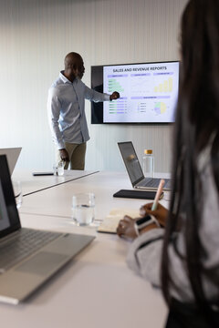 African American coworkers presenting sales charts on wall display at meeting table with laptops
