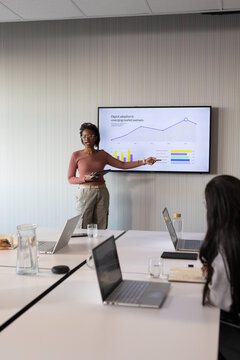 Diverse team watching woman wearing rose top presenting data on screen, holding tablet in boardroom