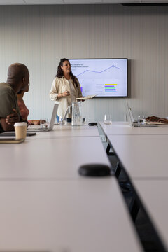 Diverse team listening while Indian woman presenting in conference room with notebook and screen