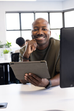 African American man leaning on desk holding tablet in folio at office, wearing olive-green top